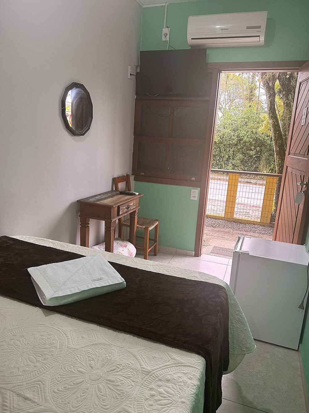 Cama da Suíte da Pousada Capricórnio vista do lado oposto, com TV, ar-condicionado e porta para a área externa