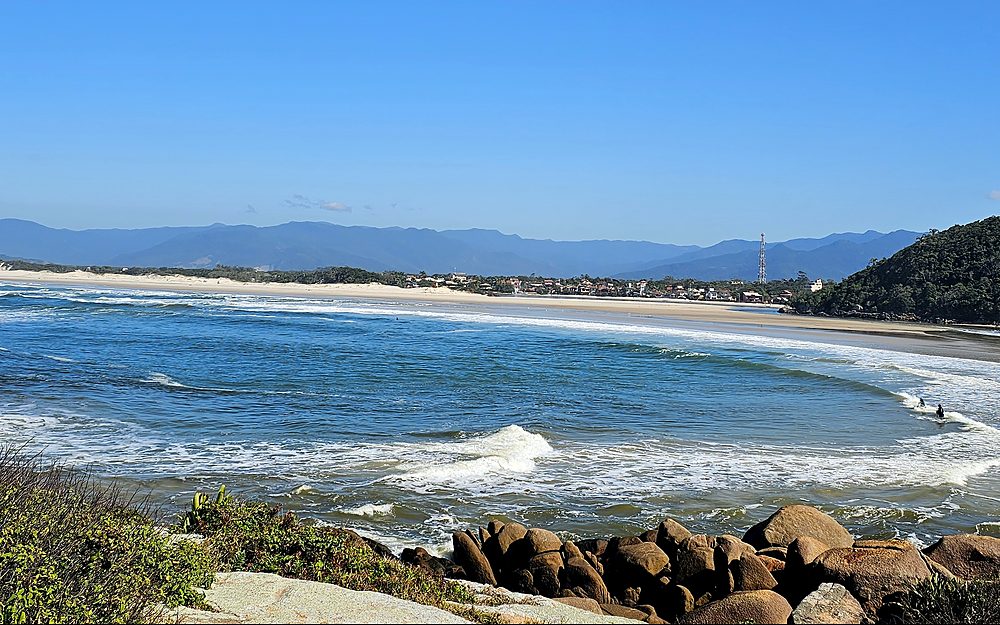Ondas quebrando ao longo da praia da Guarda do Embaú, com a vila e a serra ao fundo